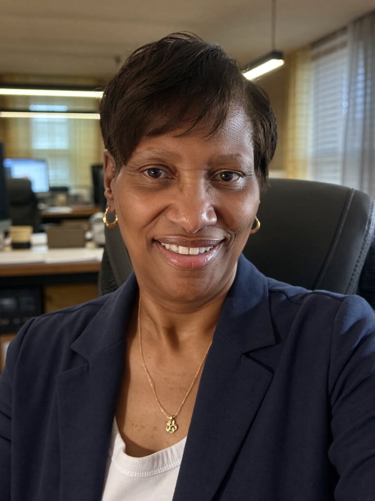 Professional woman smiling at camera in office setting, wearing dark blazer and pendant necklace